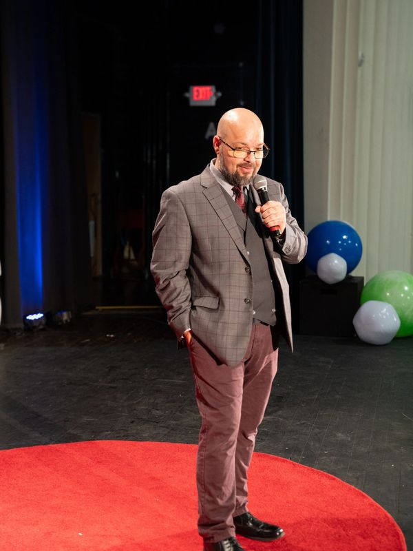 Man in glasses speaking on stage with a microphone at a red carpet spot.