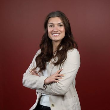 A young lady with long brown hair wearing ivory blazer, sitting with arms crossed, smiling.