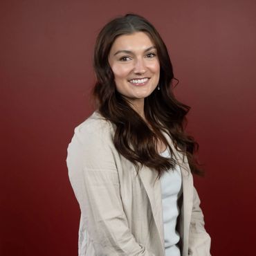 A young lady with long brown hair wearing ivory blazer & white shirt, sitting, smiling at camera.