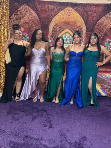 Five women in elegant evening gowns posing together indoors.