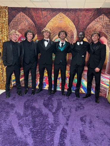 Six young men in tuxedos posing on a purple carpet with a colorful backdrop.