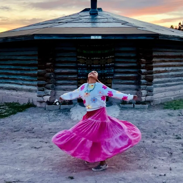 Woman spinning joyfully in front of a rustic wooden house at sunset.