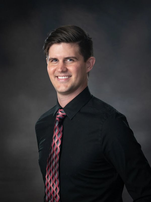 Young man in black shirt and patterned tie smiling.