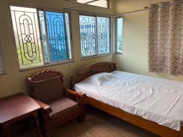 A simple bedroom with a wooden bed, chair, and side table near large windows.