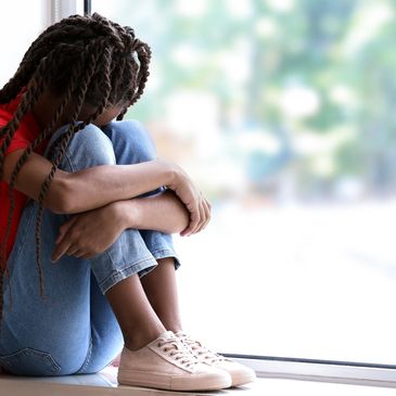 Sad Black girl with braids curled up in a window with head down and arms wrapped around her knees
