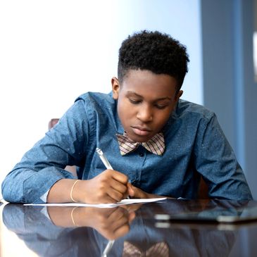 Young Black male writing at a table wearing a denim shirt and bow tie