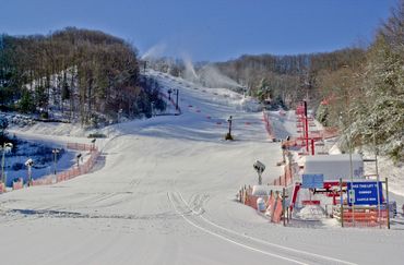 Snow-covered ski slope with chairlift and snowmaking machines under clear blue sky.