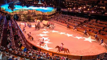 A lively indoor rodeo show with horses, riders, and a covered wagon in a large arena.