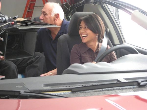 Image of a woman smiling sitting in a car at a production set. A camera and man is seen in the backg