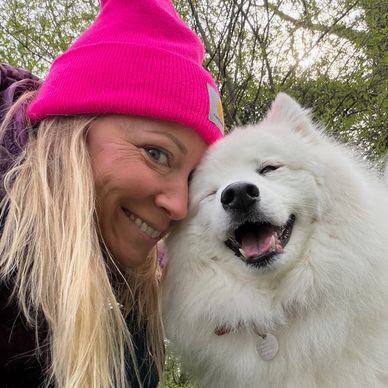 Woman in pink hat happily posing with a smiling fluffy white dog outdoors.