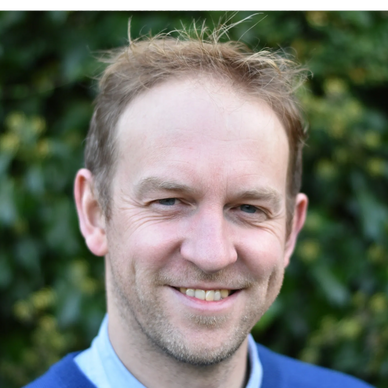 Smiling man with short hair outdoors against green foliage.