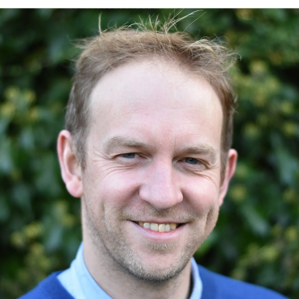 Smiling man with short hair outdoors against green foliage.