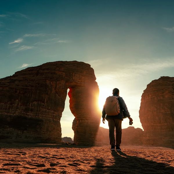 A traveler with a backpack walks towards a natural rock arch at sunset.