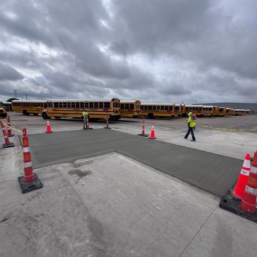 Workers in safety vests working on a concrete pavement near school buses under cloudy sky.