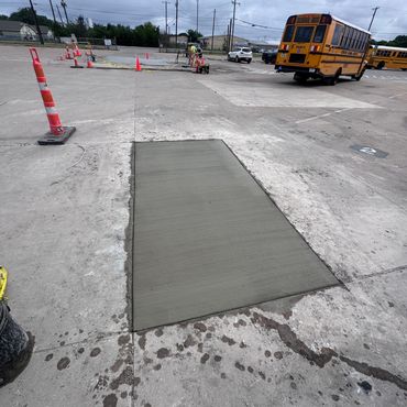 Freshly poured rectangular concrete patch on a parking lot with school buses in background.