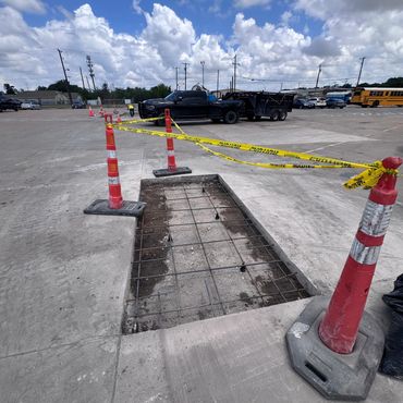 Construction site with a rectangular hole in concrete surrounded by safety cones and caution tape.