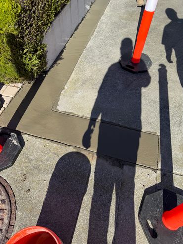 Shadows of people and cones on a sidewalk with fresh concrete patch.