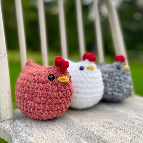 Three crocheted chickens in red, white, and gray sitting on a wooden chair.