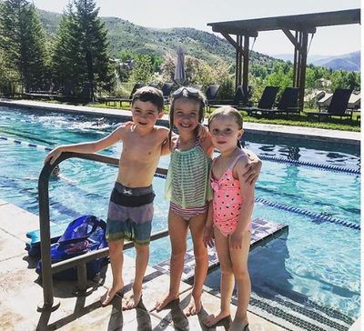 Three children in swimsuits posing by a pool on a sunny day with mountains in the background.