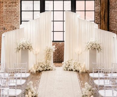 Elegant indoor wedding aisle with white floral arrangements and transparent chairs.