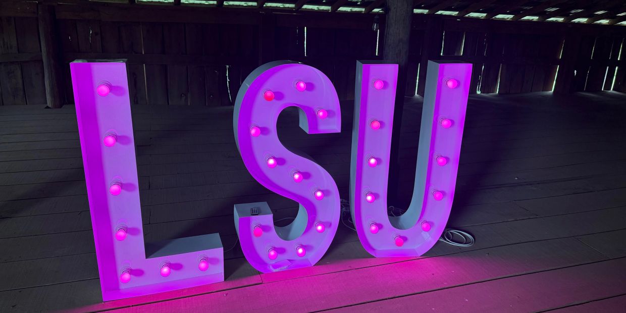 Large illuminated purple letters L, S, and U on a wooden floor.