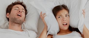 Woman covering ears with pillows next to a snoring man in bed.