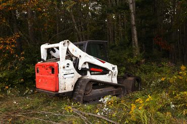 A compact skid steer loader in a forest clearing with autumn foliage.