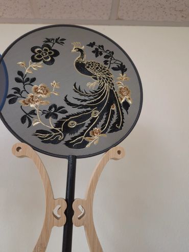 Embroidered fan with a black and gold peacock on a wooden stand.