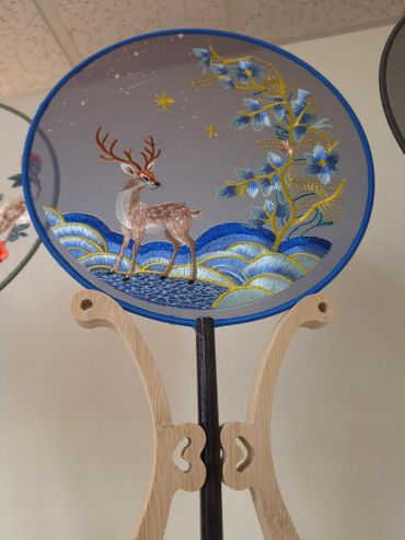 Embroidery fan with a deer and floral design on a wooden stand.