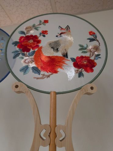 Intricately embroidered fox and floral fan on wooden stand.