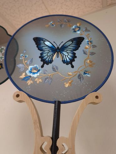 Round fan with blue butterfly and floral embroidery on a wooden stand.