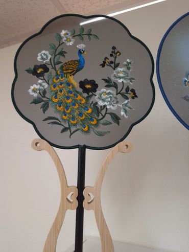 Decorative embroidered fan with peacock and floral design on wooden stand.