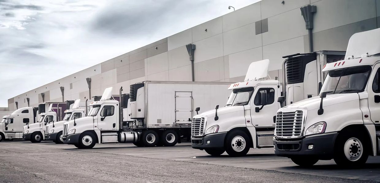 A lineup of white semi-trucks parked outside a warehouse under a cloudy sky.