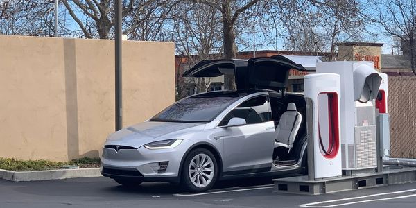 Image of a silver Tesla model X parked at a charger with the falcon wing doors open.