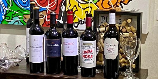 Five bottles of red wine lined up on a table with two wine glasses.