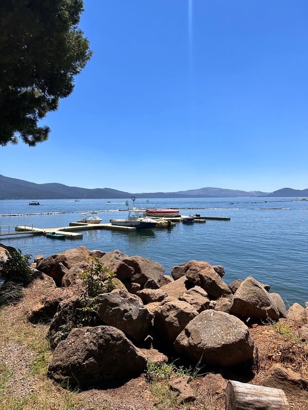 Sunny lakeside view with boats docked and mountains in the background.