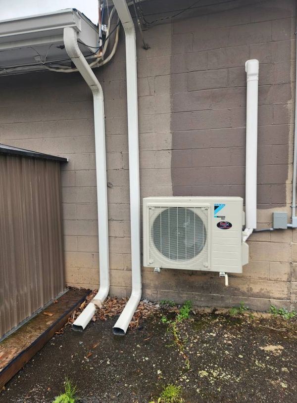 Outdoor air conditioning unit mounted on a wall with two white downspouts beside it.