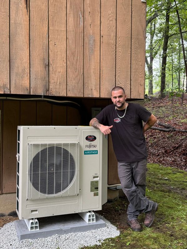 Technician proudly poses next to a newly installed Fujitsu air conditioning unit outside a wooden house.