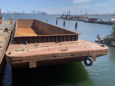 Empty flat-deck barge docked at a harbor with cranes in the distance.
