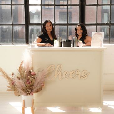 Two women smiling behind a white bar counter with "Cheers" sign and floral decoration.