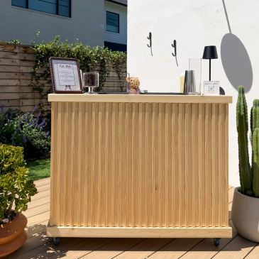 Minimalist outdoor wooden bar setup on a sunny deck with plants.