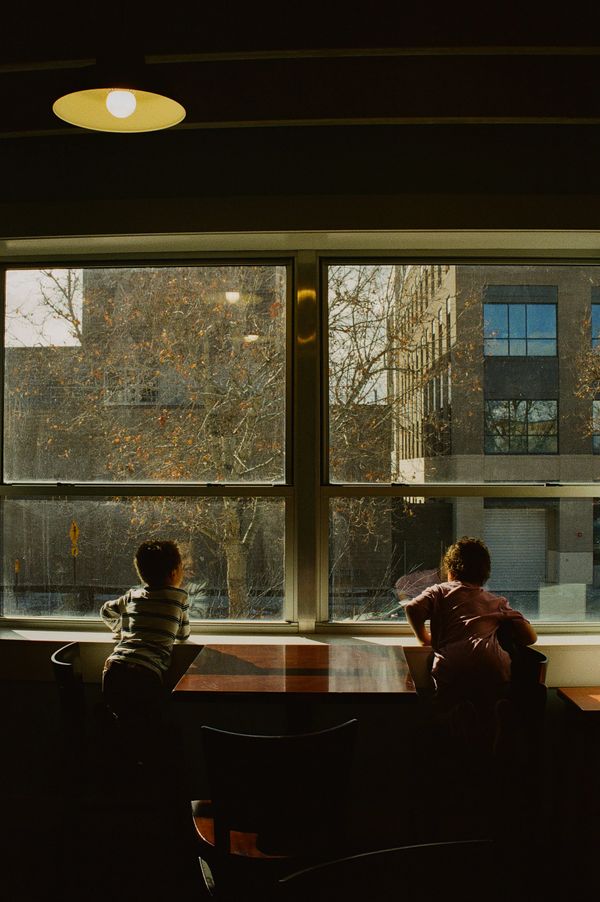 Two children looking out a large window in a dim room.