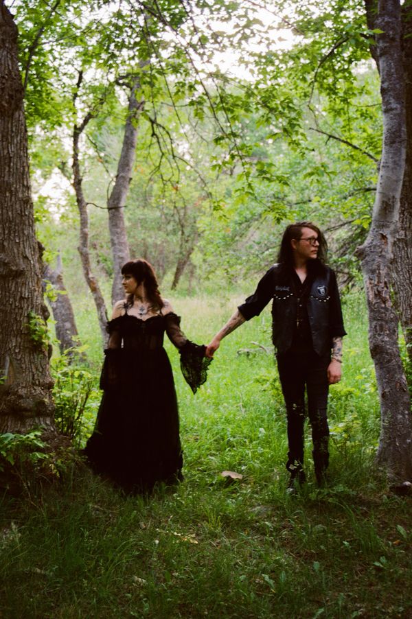 Couple dressed in gothic attire holding hands in a lush green forest.