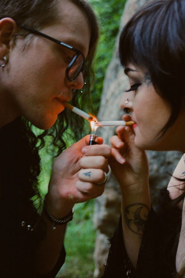 Two people with tattoos lighting cigarettes together outdoors.