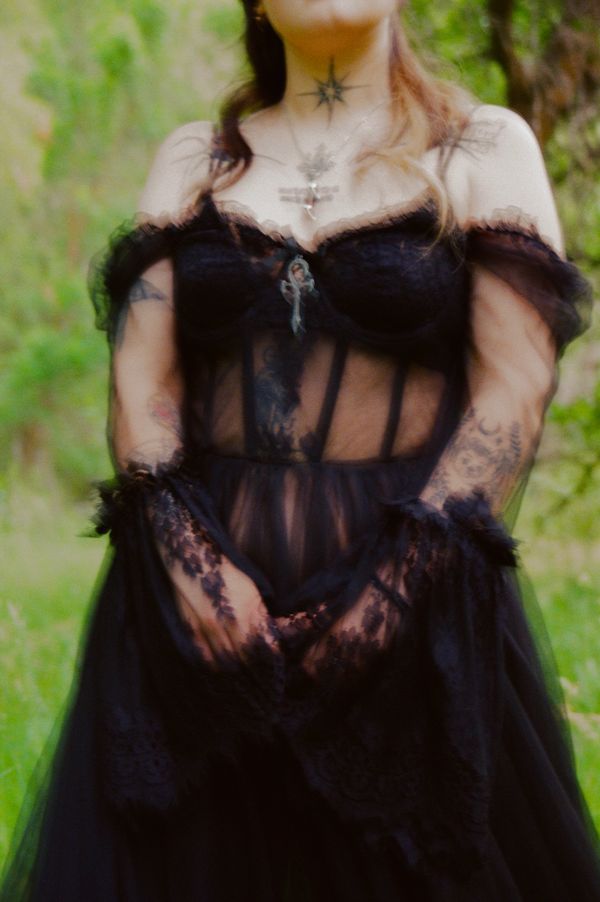 Person in a sheer black lace dress with tattoos and necklaces in a green outdoor setting.