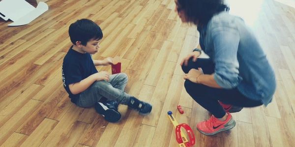 Child sitting on floor playing with toys, adult crouching nearby.