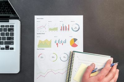 Person holding pen and notebook near laptop and charts on desk.