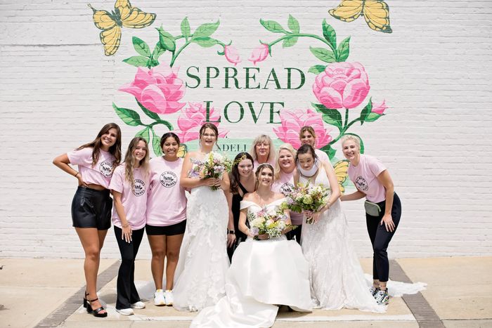 Group of 11 women standing in front of a mural reading "Spread Love." 3 women are in wedding dresses