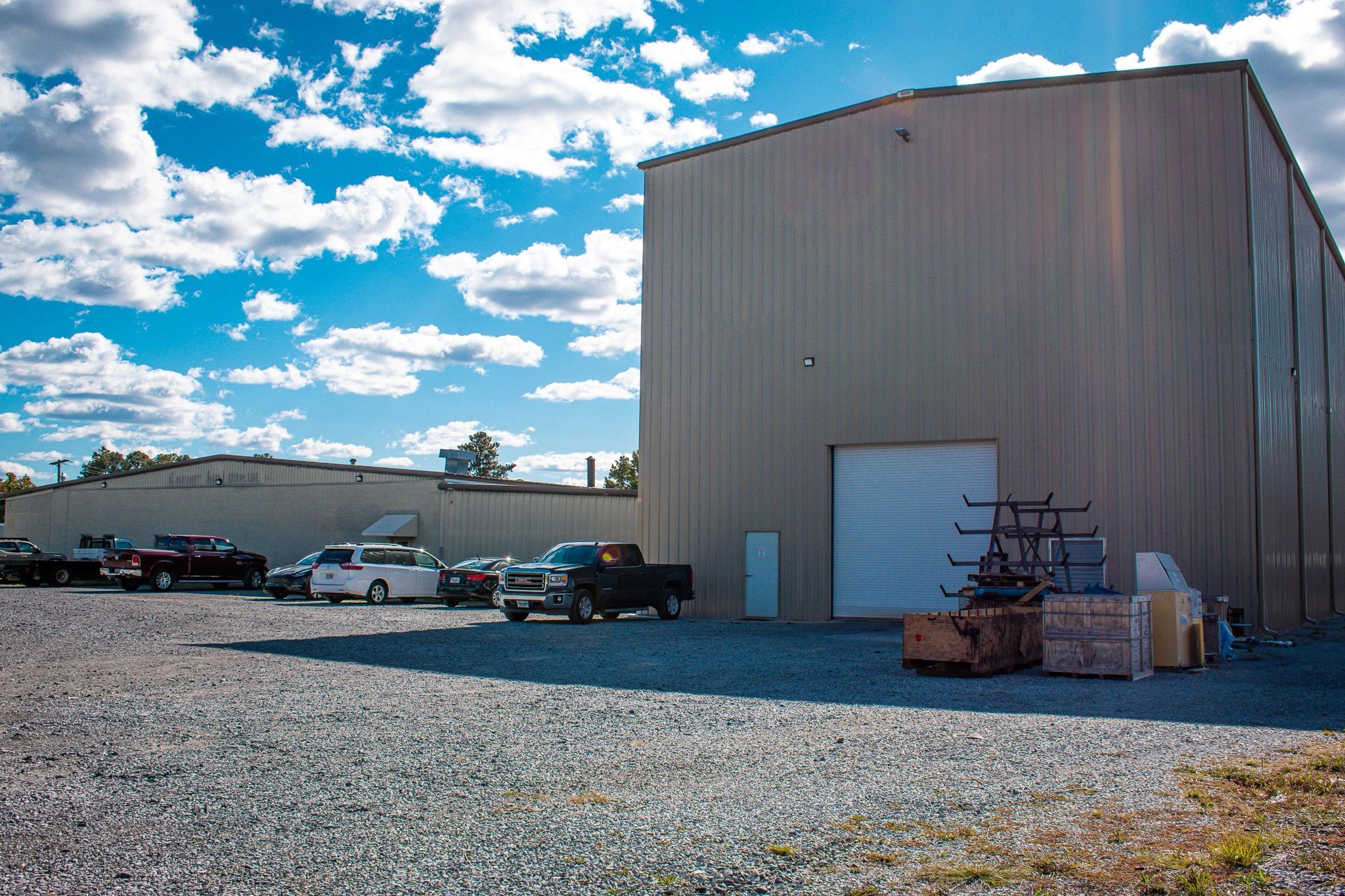 Metal building and high bay with cars parked in front. Bright blue sky and clouds in background.