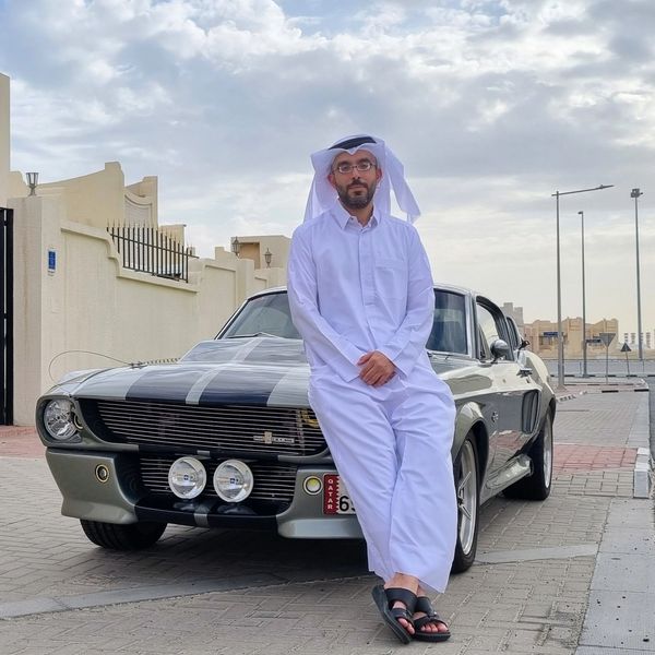 Man in traditional attire leaning on a classic car outdoors.